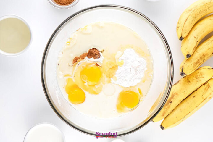A glass bowl with eggs, flour, cinnamon, and milk surrounded by bananas and small bowls of ingredients on a white surface.