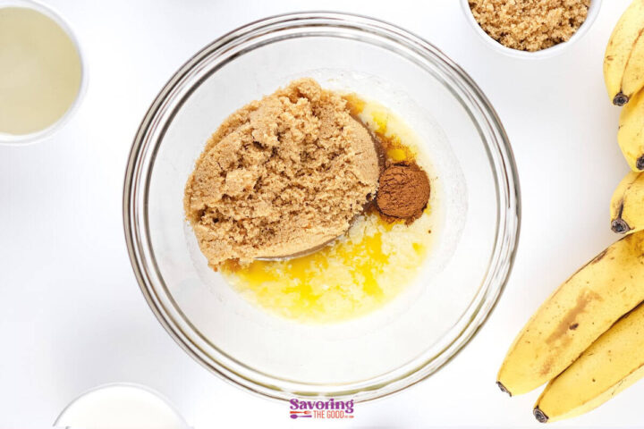A glass bowl with brown sugar, melted butter, and cinnamon surrounded by bananas and other baking ingredients on a white surface.
