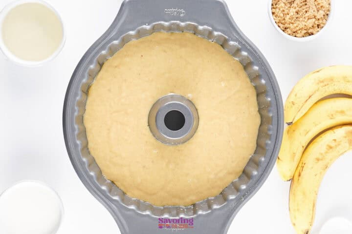 A bundt pan filled with banana cake batter is surrounded by bananas, brown sugar, oil, and a white liquid in small bowls, ready for baking.