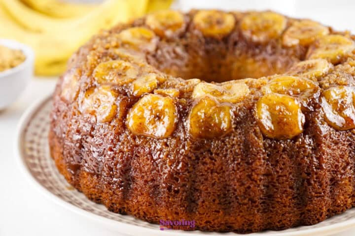 A close-up of a glazed bundt cake topped with caramelized banana slices, displayed on a white plate.