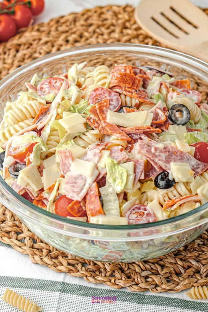 A glass bowl filled with pasta salad containing rotini, sliced meats, cheese, black olives, grape tomatoes, and lettuce, placed on a woven placemat.