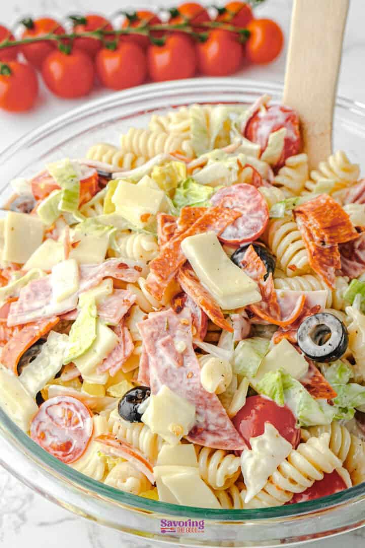 A glass bowl filled with pasta salad containing rotini, sliced pepperoni, salami, cheese cubes, black olives, lettuce, and cherry tomatoes, with a wooden spoon in the bowl.