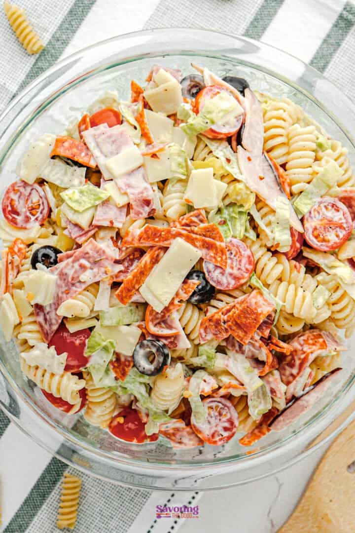 A glass bowl filled with Italian pasta salad containing rotini pasta, cherry tomatoes, olives, lettuce, sliced meats, cheese, and a creamy dressing.