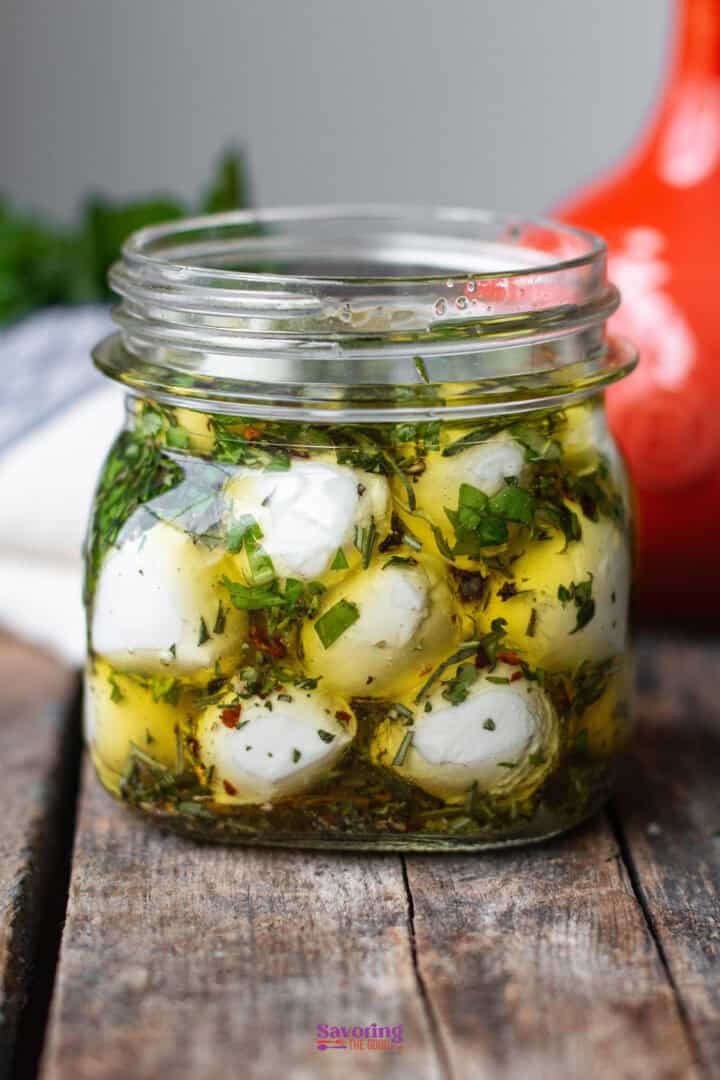 A glass jar filled with marinated mozzarella balls in olive oil, herbs, and spices, sits on a wooden surface.