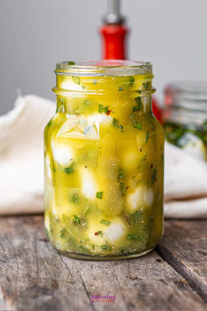A glass jar filled with marinated mozzarella balls, herbs, and oil sits on a wooden surface with a blurred background.