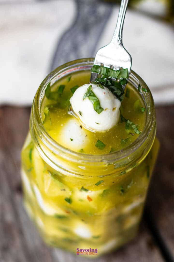 A fork lifts a mozzarella ball out of a jar filled with marinated mozzarella balls, herbs, and oil.