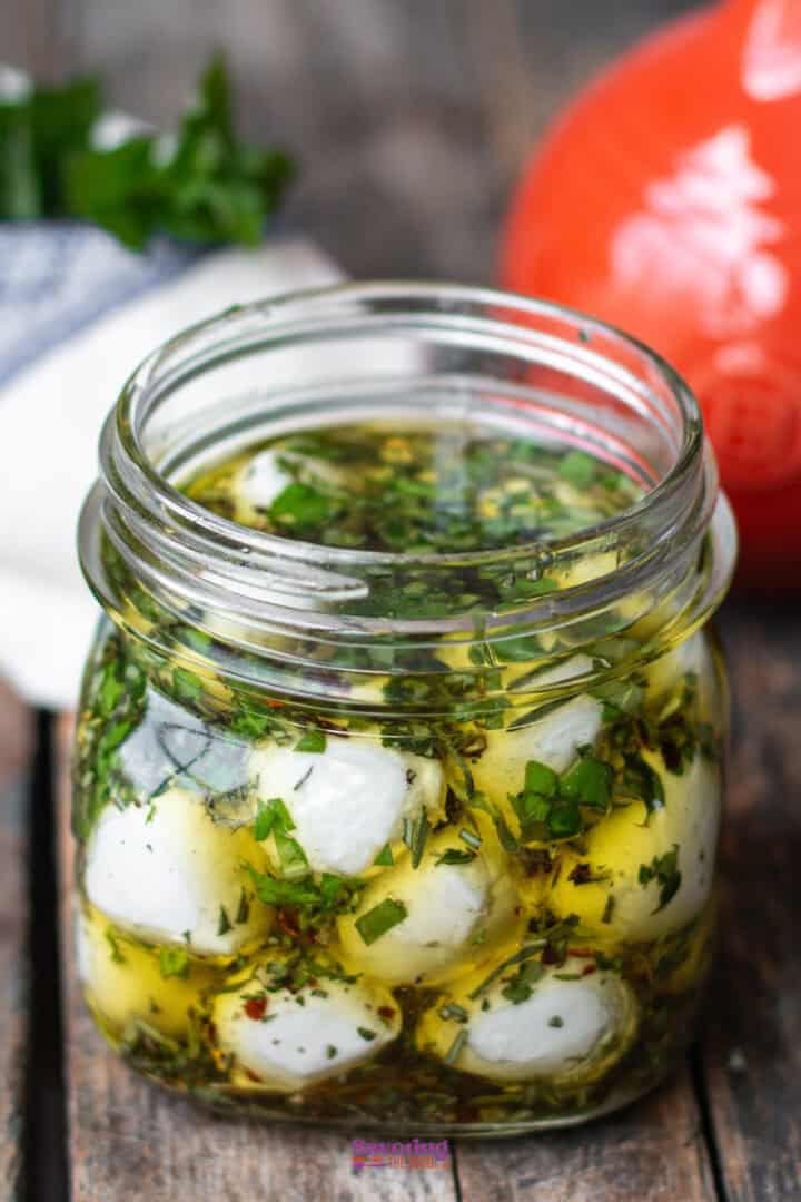 A glass jar filled with marinated mozzarella balls in olive oil with herbs, placed on a wooden surface.