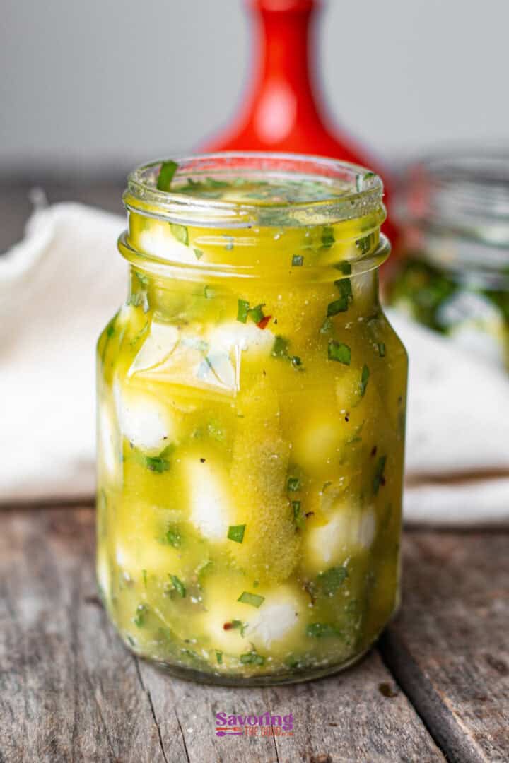 A jar filled with marinated cheese cubes in a herbed oil mixture on a wooden table, with a red object and a white cloth in the background.