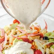 A measuring cup pours Antipasto Pasta Salad dressing onto a bowl of pasta salad with rotini, lettuce, cherry tomatoes, black olives, and sliced deli meats.