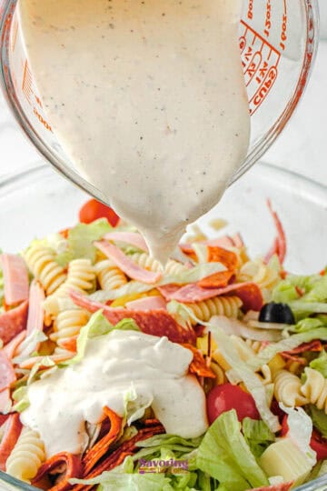 A measuring cup pours Antipasto Pasta Salad dressing onto a bowl of pasta salad with rotini, lettuce, cherry tomatoes, black olives, and sliced deli meats.