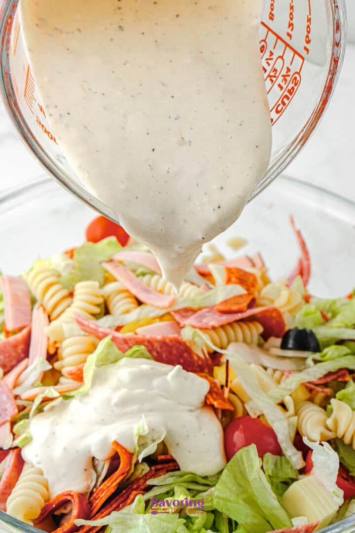 A measuring cup pours Antipasto Pasta Salad dressing onto a bowl of pasta salad with rotini, lettuce, cherry tomatoes, black olives, and sliced deli meats.
