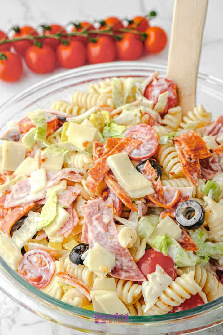 A glass bowl filled with Antipasto Pasta Salad featuring rotini, sliced pepperoni, salami, cheese, black olives, lettuce, and cherry tomatoes, served with a wooden spoon.