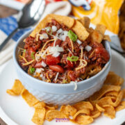 A bowl of chili pie topped with shredded cheese, chopped onions, and green onions, surrounded by crunchy Fritos corn chips on a white plate.