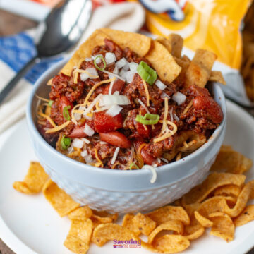 A bowl of chili pie topped with shredded cheese, chopped onions, and green onions, surrounded by crunchy Fritos corn chips on a white plate.
