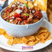 A bowl of chili pie topped with diced onions, shredded cheese, and green onions, served with Fritos corn chips on a white plate on a wooden table.