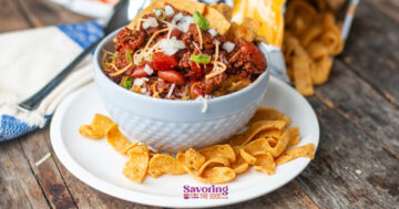 A bowl of chili pie topped with diced onions, shredded cheese, and green onions, served with Fritos corn chips on a white plate on a wooden table.