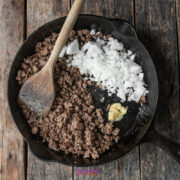 Ground beef, chopped onions, and minced garlic cooking in a cast iron skillet on a wooden surface, with a wooden spoon resting in the pan.