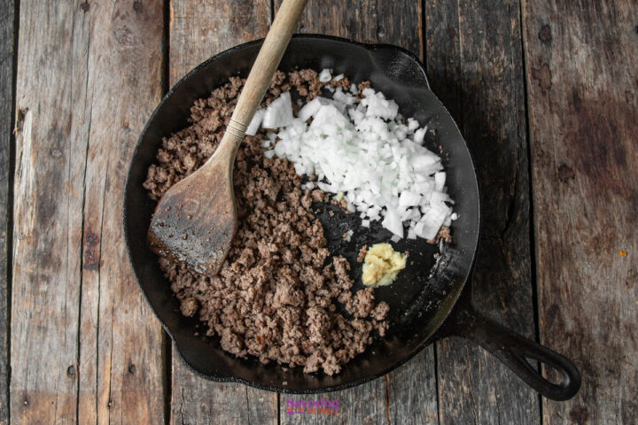 Ground beef, chopped onions, and minced garlic cooking in a cast iron skillet on a wooden surface, with a wooden spoon resting in the pan.
