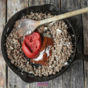 A cast iron skillet with cooked ground beef, diced onions, tomato paste, spices, and a wooden spoon on a rustic wooden surface.