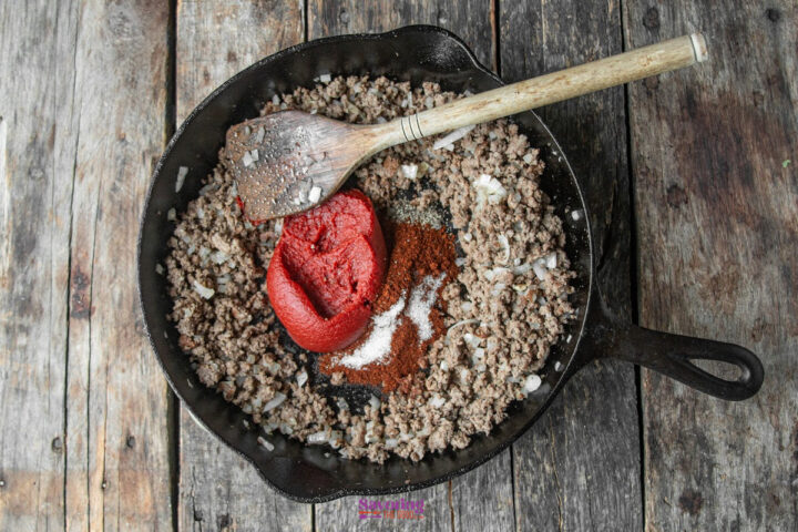 A cast iron skillet with cooked ground beef, diced onions, tomato paste, spices, and a wooden spoon on a rustic wooden surface.