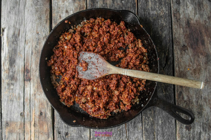 A cast iron skillet filled with cooked ground meat and sauce, with a wooden spoon resting inside, sits on a weathered wooden surface.