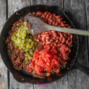 A cast iron skillet on a wooden surface filled with cooked ground meat, diced green chiles, kidney beans, and chopped tomatoes being mixed with a wooden spoon.