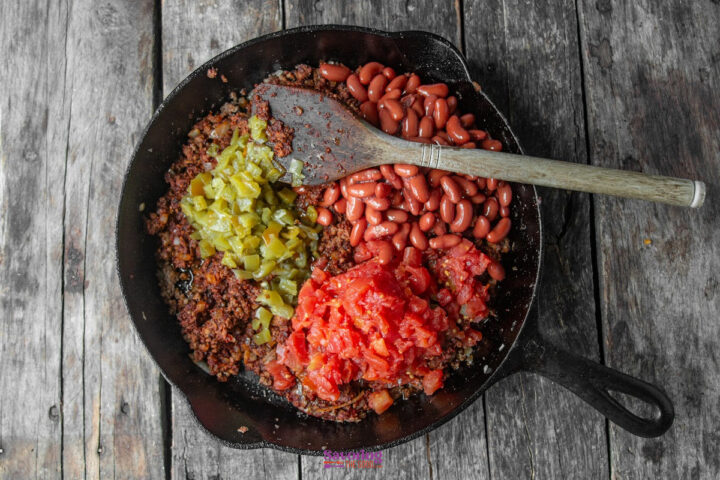 A cast iron skillet on a wooden surface filled with cooked ground meat, diced green chiles, kidney beans, and chopped tomatoes being mixed with a wooden spoon.