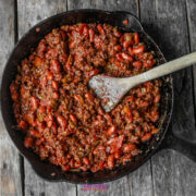 A cast iron skillet filled with chili made of ground beef, kidney beans, and tomato sauce, with a wooden spoon resting inside, on a rustic wooden surface.