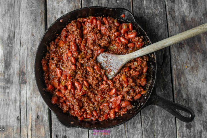 A cast iron skillet filled with chili made of ground beef, kidney beans, and tomato sauce, with a wooden spoon resting inside, on a rustic wooden surface.