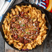 A cast iron skillet filled with chili pie fritos, topped with melted cheese, chopped onions, and Fritos corn chips, with a Fritos bag and a cloth napkin nearby on a wooden table.