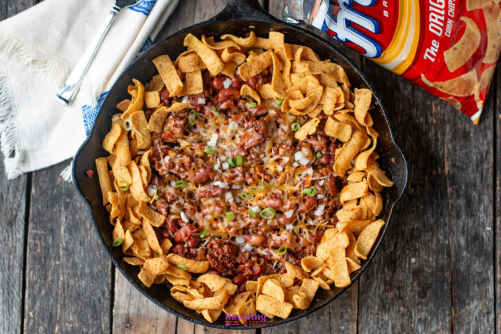 A cast iron skillet filled with chili pie fritos, topped with melted cheese, chopped onions, and Fritos corn chips, with a Fritos bag and a cloth napkin nearby on a wooden table.