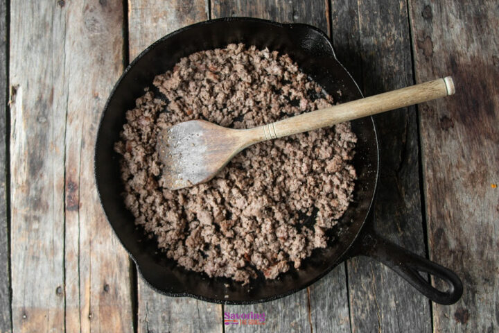 Cooked ground beef in a cast iron skillet with a wooden spoon, ready for your next chili pie Fritos creation, placed on a rustic wooden surface.