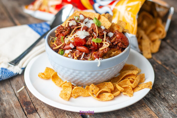A bowl of chili pie topped with onions, cheese, and Fritos chips sits on a plate surrounded by more Fritos, with a spoon and a partially opened chip bag in the background.