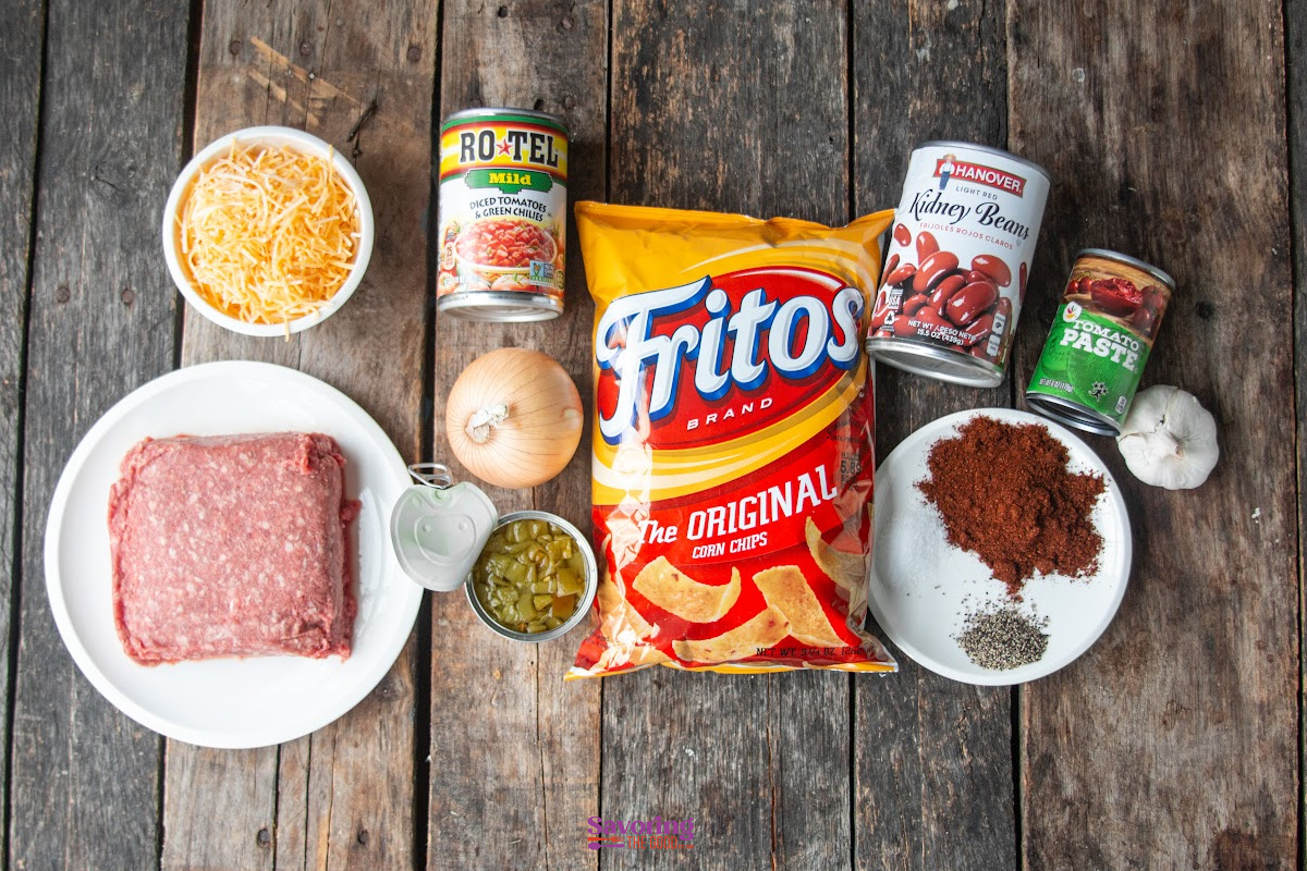 Ingredients for a chili pie fritos dish arranged on a wooden surface, including ground beef, cheese, Fritos, beans, Rotel, tomato paste, onion, garlic, spices, and chopped jalapeños.