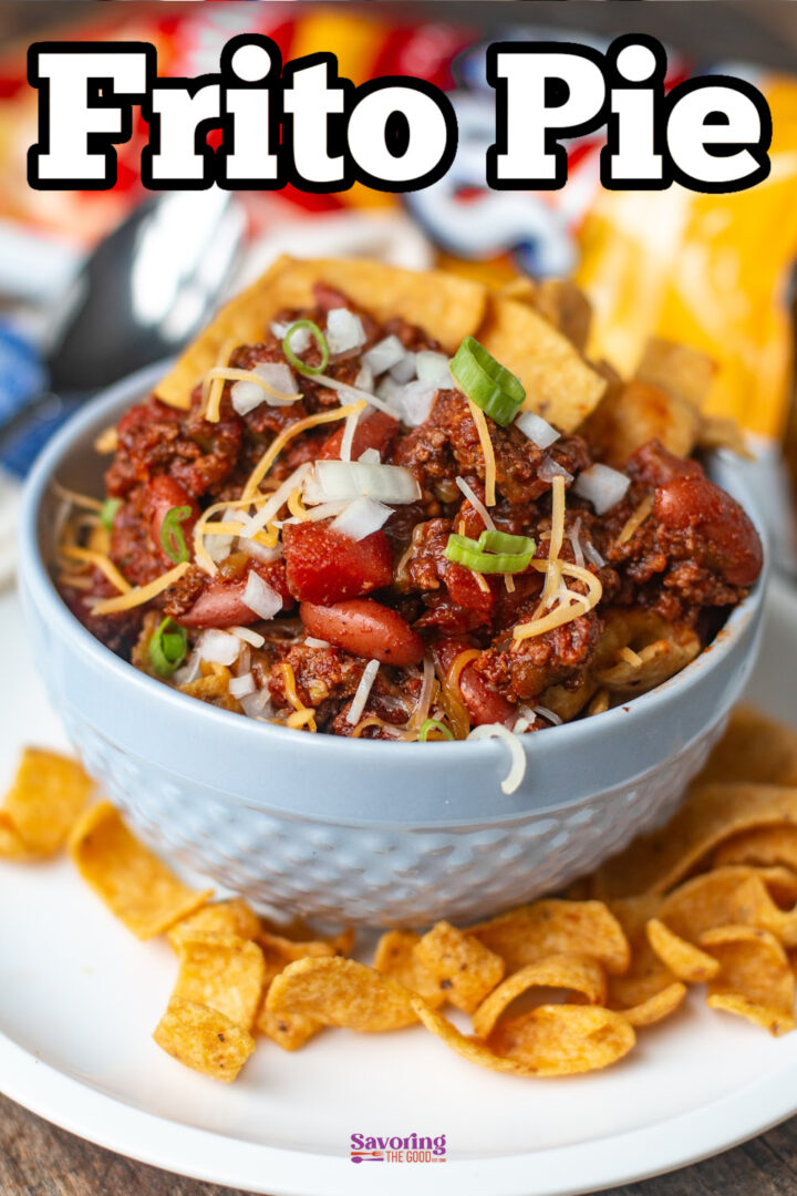 A bowl of Frito pie topped with chili, shredded cheese, chopped onions, and green onions, with corn chips scattered around the bowl on a plate.
