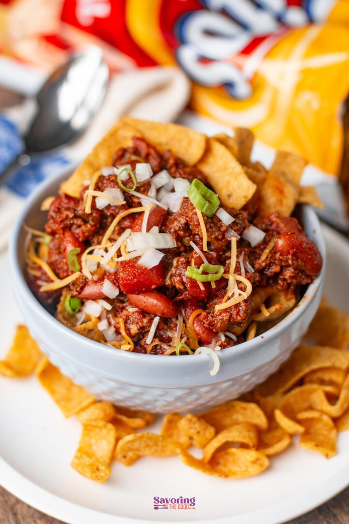 A bowl of chili topped with shredded cheese, chopped onions, and green onions, served with corn chips on the side.