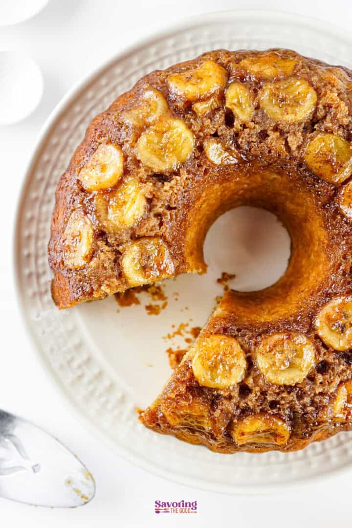 A ring-shaped caramelized banana cake with a slice removed, displayed on a white textured plate.