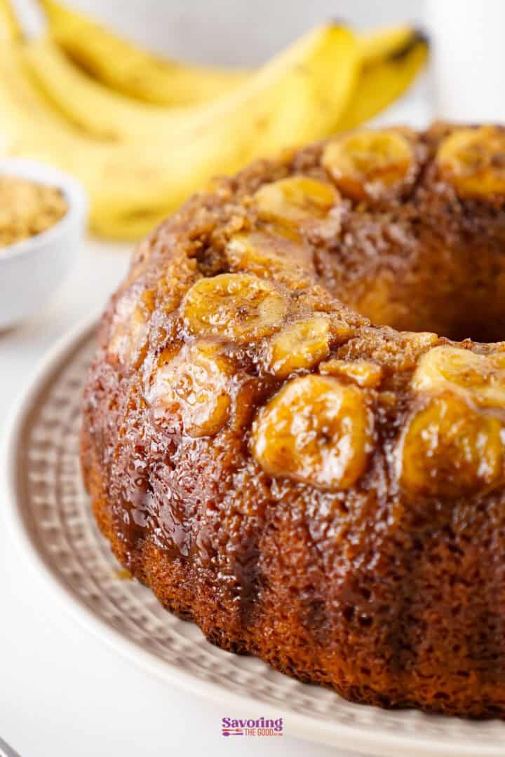 A banana upside-down cake on a plate, topped with caramelized banana slices. Bananas and a bowl of chopped nuts are visible in the background.