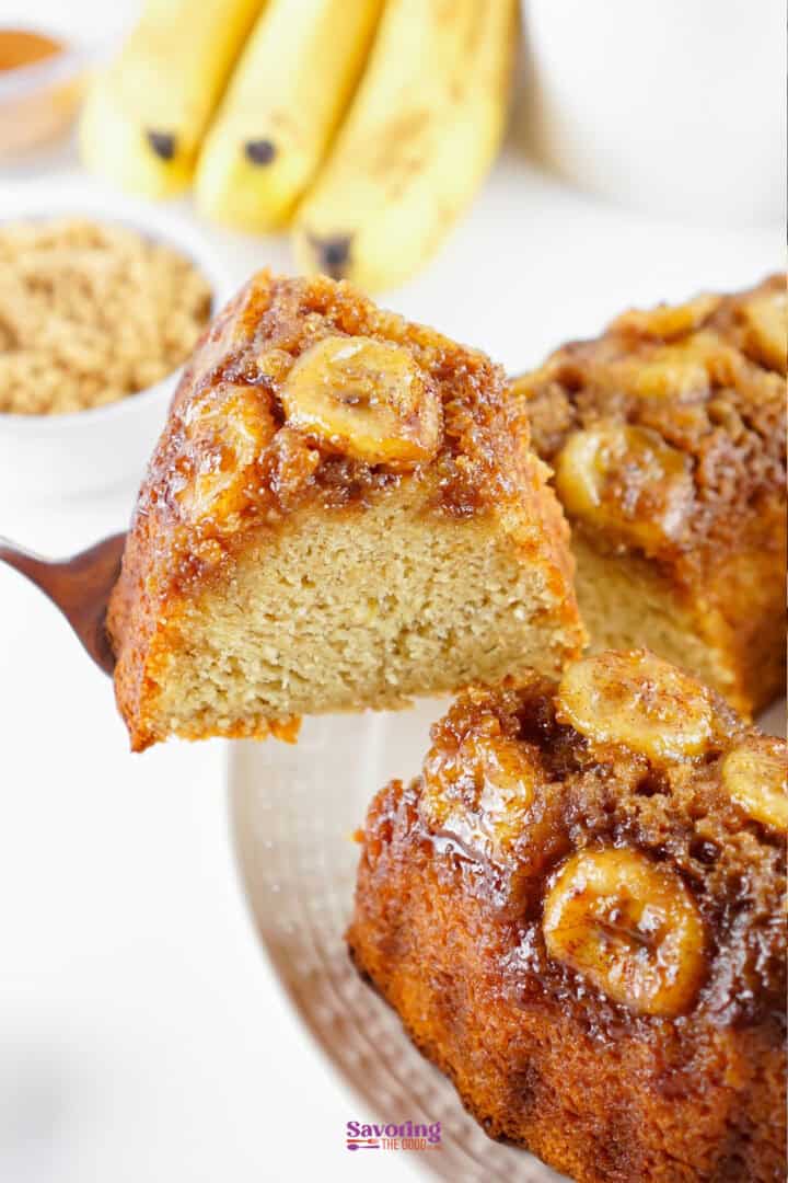 A slice of banana upside-down cake with caramelized banana topping is being lifted from a plate; bananas and brown sugar are in the background.