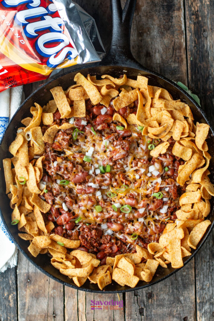 A cast iron skillet with chili topped with shredded cheese, chopped onions, green onions, and Fritos corn chips arranged around the edge.