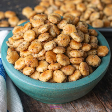 A teal bowl filled with seasoned oyster crackers sits on a wooden table, with more crackers scattered in the background.