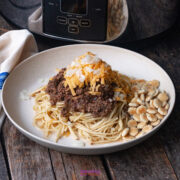 A plate of spaghetti topped with Cincinnati chili, shredded cheddar cheese, chopped onions, and a side of oyster crackers sits on a wooden table near a crockpot.