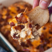 A hand holds a slice of bread topped with a cheesy caramelized onion dip with bacon, with more of the savory dip visible in a dish in the background.