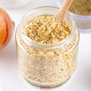 A glass jar filled with Onion Soup Mix, with a wooden spoon scooping some out; an onion sits nearby.