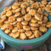 A bowl filled with seasoned oyster crackers sits on a wooden surface next to a white and blue towel, with more crackers on a tray in the background.