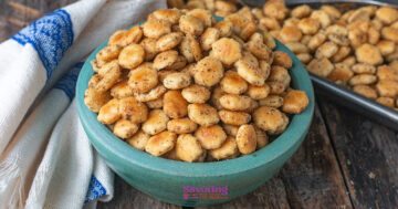 A bowl filled with seasoned oyster crackers sits on a wooden surface next to a white and blue towel, with more crackers on a tray in the background.