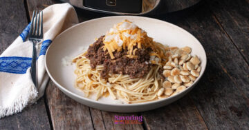A plate of spaghetti topped with chili, shredded cheese, and diced onions, served with oyster crackers on the side.
