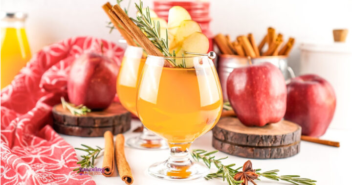 Two glasses of apple cider with Fireball, garnished with apple slices and rosemary, are surrounded by red apples, cinnamon sticks, and a red cloth.