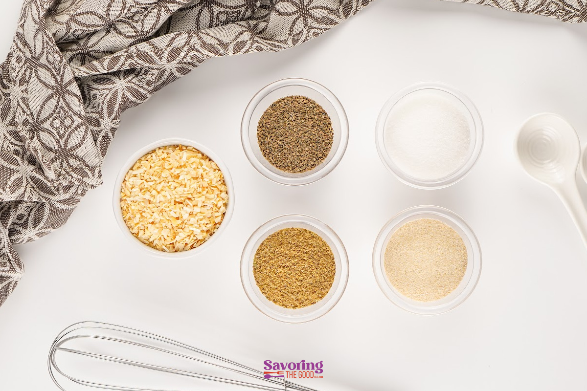 Five small containers hold dried minced onion, black pepper, salt, celery seed, and garlic powder-classic ingredients for a homemade Onion Soup Mix-arranged on a white surface with a patterned cloth, spoon, and whisk nearby.