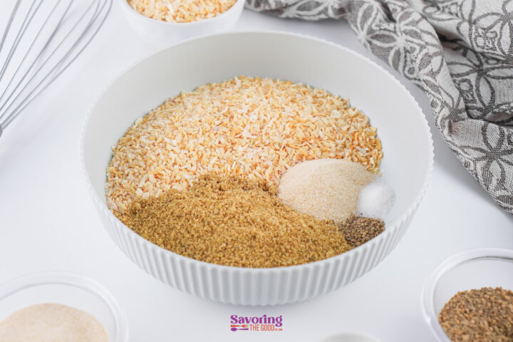 A white bowl filled with dried minced onion, onion powder, garlic powder, salt, pepper, and other spices-perfect for homemade Onion Soup Mix-sits on a white surface with a gray patterned cloth nearby.